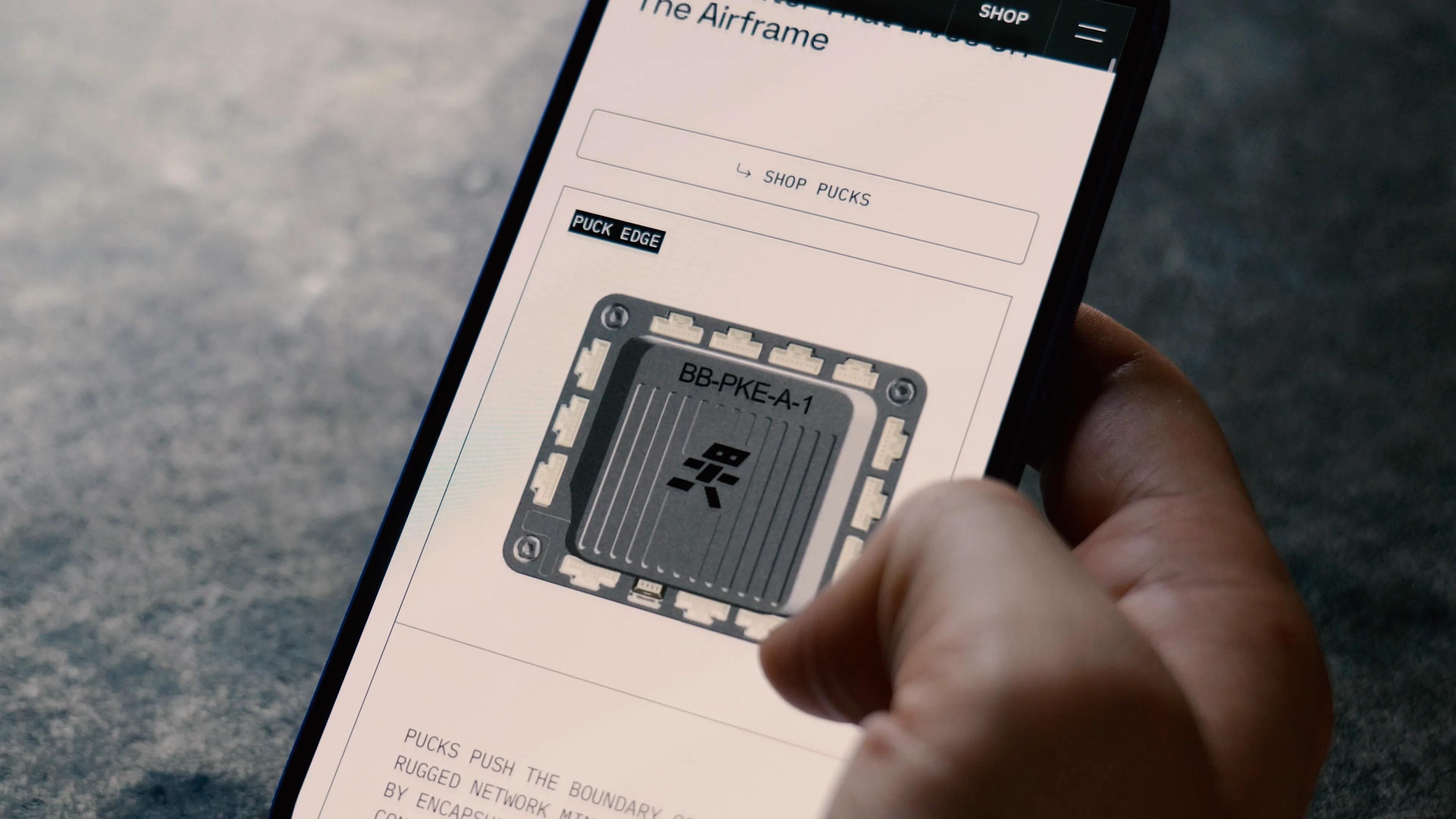 A hand holding a phone that shows a 3D render of the BotBlox Puck Edge networking module on its product page