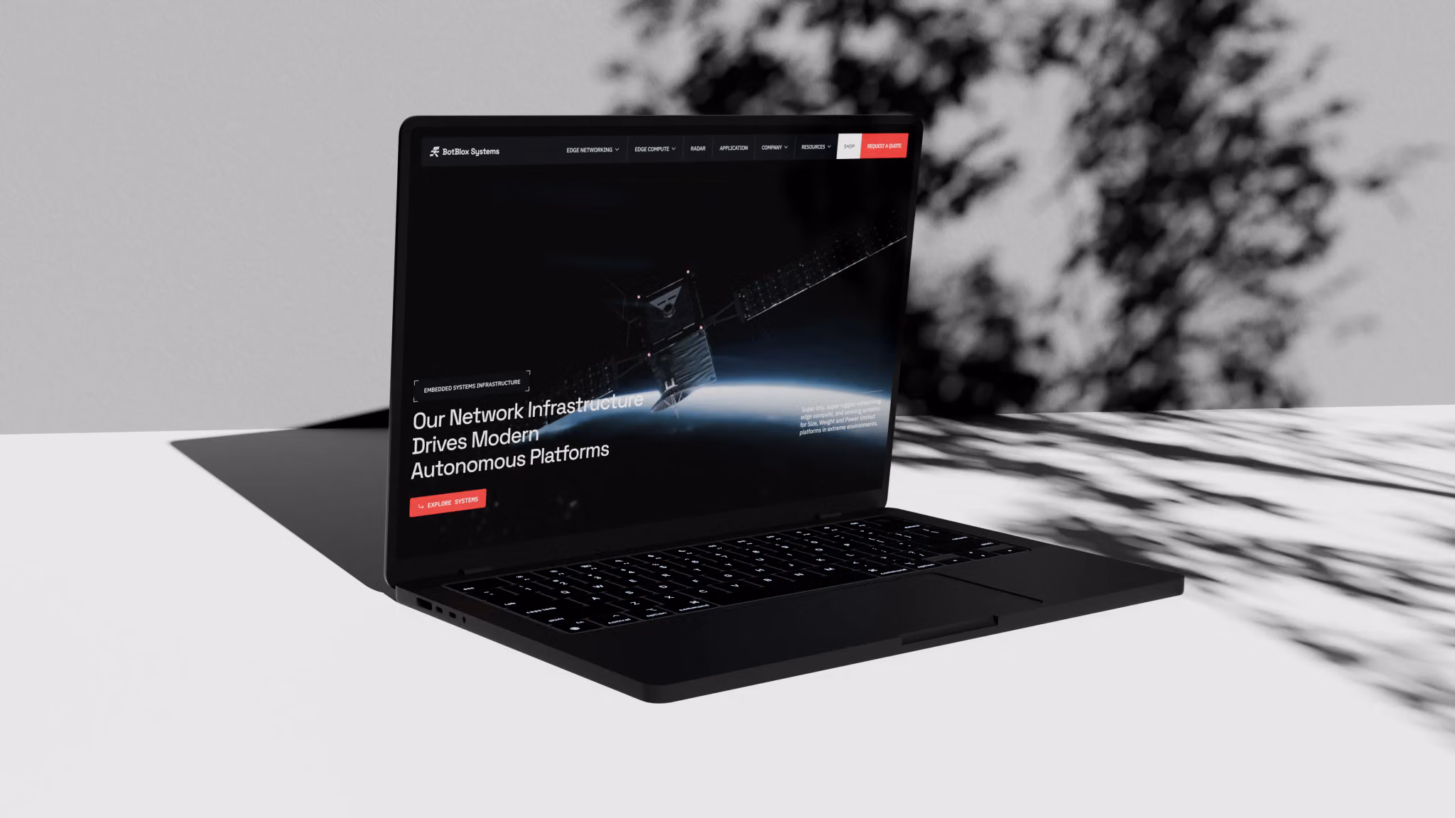 The BotBlox Systems website on a laptop in dappled tree shade: a satellite above Earth and a headline on autonomous platforms